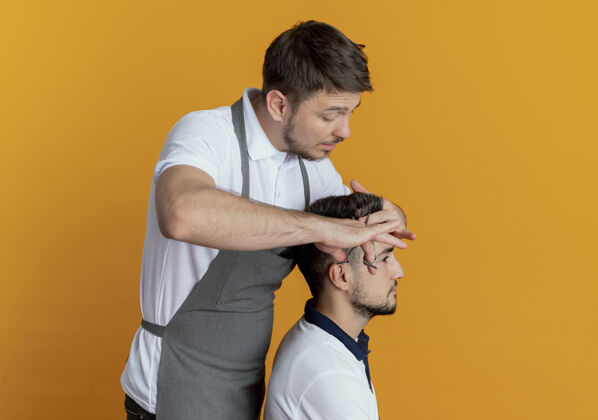 男人穿着围裙的理发师用满意顾客的剪刀在橙色的墙上剪头发满意剪刀围裙