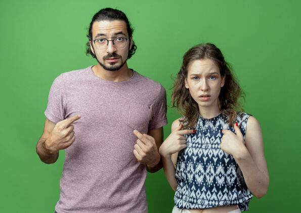 愤愤不平愤怒的年轻夫妇指着自己站在绿墙旁边年轻人男人指着