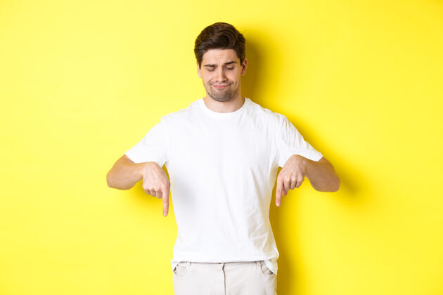 秀穿着白色t恤的怀疑的年轻人 指着并低头沮丧 不赞成和不喜欢的产品 站在黄色的背景下年轻时尚男人