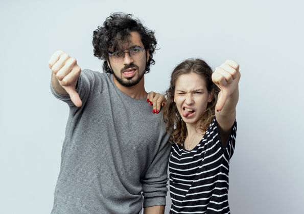 拇指年轻漂亮的夫妇不高兴地在白墙上竖起大拇指向下年轻人夫妇