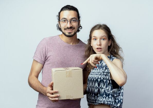 女人年轻漂亮的一对男女 男人拿着盒子包微笑着 而他的女朋友用手指着盒子惊讶地翻过白色的墙壁情侣同时抱着