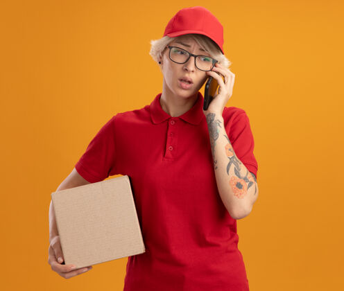 非常身穿红色制服 戴着帽子 戴着眼镜 拿着纸板箱 拿着手机的年轻送货员站在橘色的墙上 感到困惑和焦虑制服站着眼镜