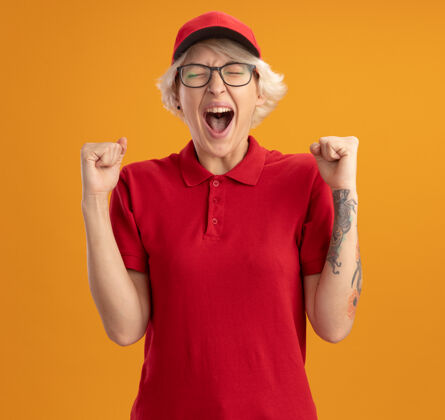 制服身穿红色制服 头戴鸭舌帽的年轻女送货员高兴而兴奋地握紧拳头 为自己的成功而欢呼雀跃 站在橙色的墙上高喊着握紧交付女人