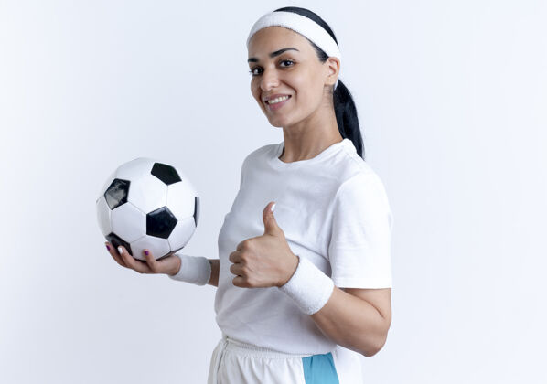 微笑年轻微笑的白人运动女性戴着头带和腕带拿着球 竖起大拇指站在一边 与空白空间隔离腕带穿年轻