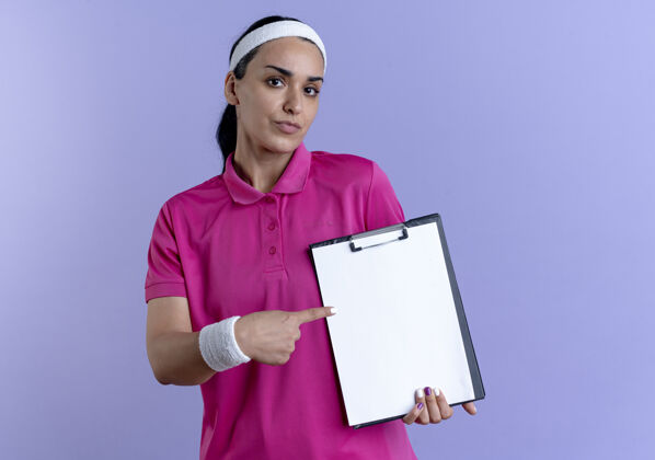 点年轻自信的高加索运动女性戴着头带和腕带 拿着并指着剪贴板 在紫色的空白处与复制空间隔离剪贴板信心年轻
