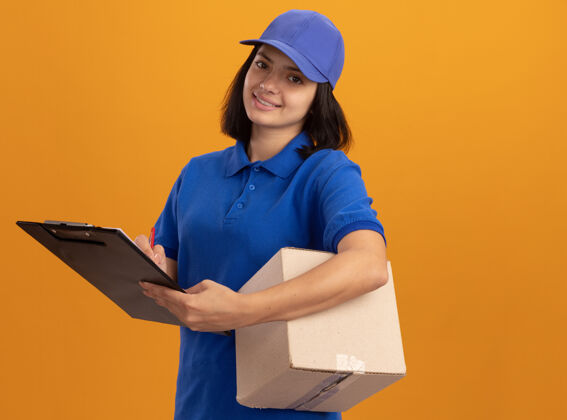 交货快乐的年轻送货女孩 穿着蓝色制服 戴着帽子 手里拿着纸板箱和剪贴板 站在橙色的墙上 脸上带着微笑脸女孩微笑
