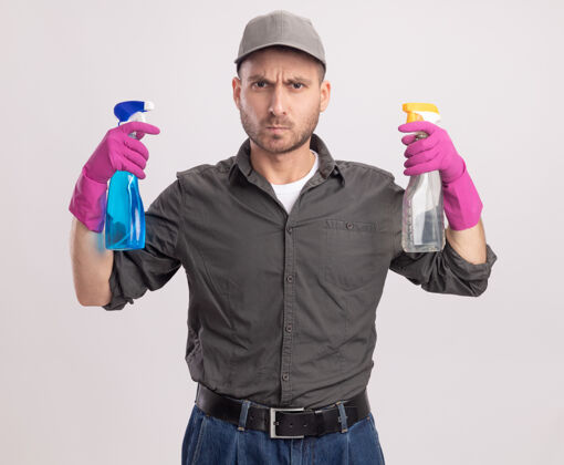 男人年轻的清洁工穿着休闲服 戴着橡胶手套 手里拿着喷雾瓶 站在白色的墙上 表情严肃休闲橡胶持有