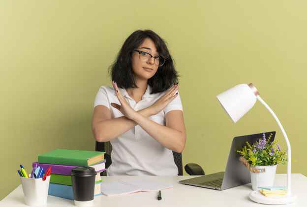 工具年轻自信漂亮的高加索女学生戴着眼镜坐在书桌旁 手里拿着学习用具 双臂交叉着 在绿色空间里用复制空间表示没有孤立手势手臂空间