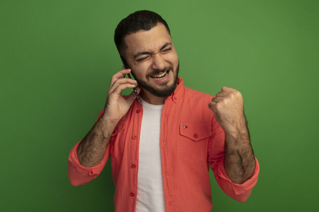 男人站在绿色的墙上 一位身穿橘色衬衫 留着胡子的年轻人一边用手机聊天 一边握紧拳头 既高兴又兴奋胡子而兴奋
