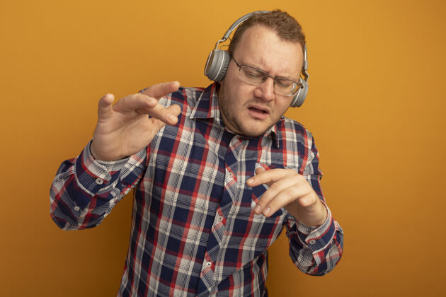 耳机戴着眼镜 穿着格子衬衫 戴着耳机的情绪化男人站在橙色的墙上欣赏音乐男人眼镜站立