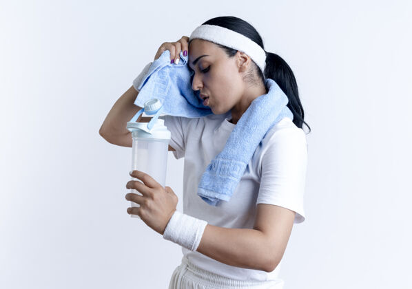 毛巾年轻疲惫的白人运动女性戴着头带和腕带站在一边 用毛巾擦拭额头拿着水瓶隔离在白色空间与复制空间复制空间运动