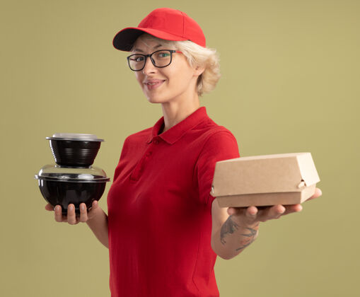 拿着年轻的送货员身穿红色制服 戴着帽子 戴着眼镜 手里拿着食品包 微笑着自信地站在绿色的墙上站着眼镜戴着