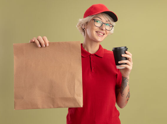 女士身穿红色制服 戴着帽子 戴着眼镜 手里拿着纸包和咖啡杯的年轻女送货员站在绿色的墙上微笑着包装眼镜微笑