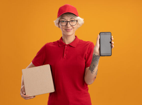 穿着身穿红色制服 头戴眼镜 手拿纸板箱 脸上挂着智能手机的年轻送货员自信地站在橙色的墙上女人微笑帽子