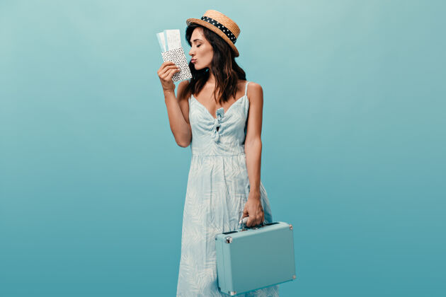 手提箱戴草帽的漂亮女士拿着蓝色背景下的时髦包和票穿着夏装摆姿势的漂亮女人肖像思考女孩