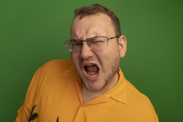 喊叫一个戴着眼镜 穿着橙色衬衫的男人站在绿色的墙上 带着恼怒的表情大喊大叫男人眼镜生气