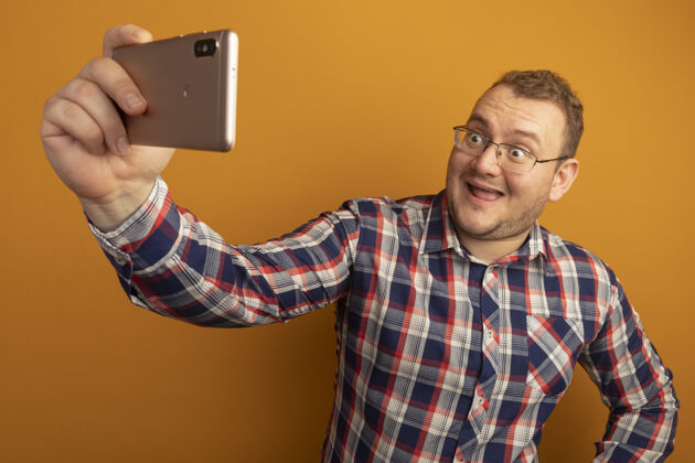 站立戴着眼镜和格子衬衫的男人用智能手机自拍 站在橘色的墙上开心地微笑着微笑使用眼镜