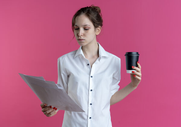 年轻年轻自信的俄罗斯金发女孩拿着咖啡杯 看着粉色空间里的纸页 还有复印空间咖啡持有床单