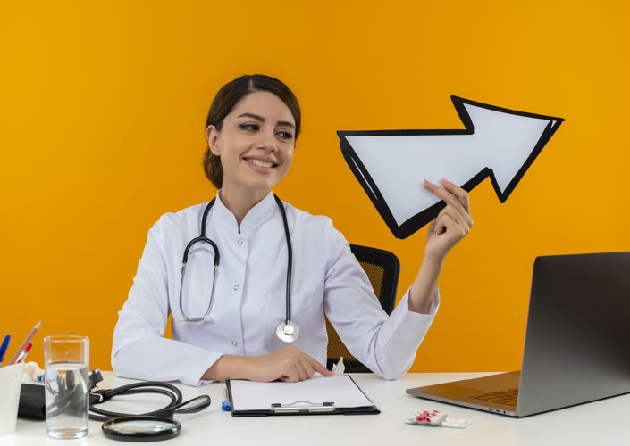 穿面带微笑的年轻女医生穿着医用长袍 手持听诊器 坐在办公桌旁 手持医疗工具和笔记本电脑 看着黄色墙壁上的箭头标记拿着笔记本电脑长袍