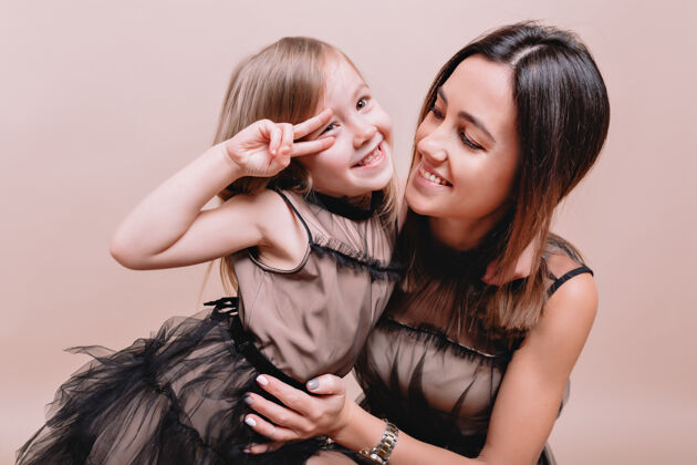 青少年迷人可爱的女孩和她时尚的母亲在米色墙上穿着相似的黑色连衣裙的特写肖像小夏天衣服