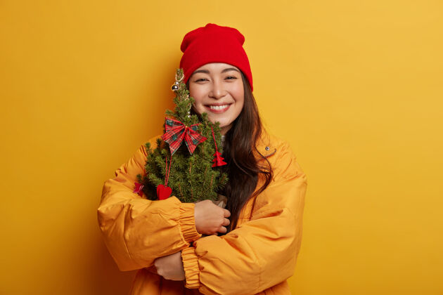 外套一位戴着红帽子 穿黄夹克的韩国女士拥抱着绿色装饰的小圣诞树 微笑着 有着喜庆的心情 孤立在黄色的背景上亚洲女人模特
