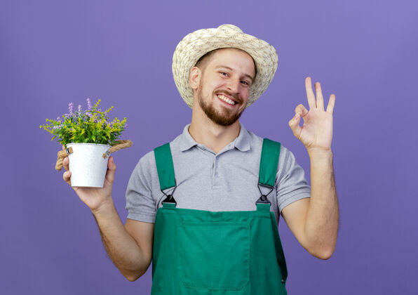 拿着面带微笑的年轻英俊的斯拉夫园丁 穿着制服 戴着帽子 手里拿着花盆 看着紫色墙上孤立的做得很好的标志年轻好看着