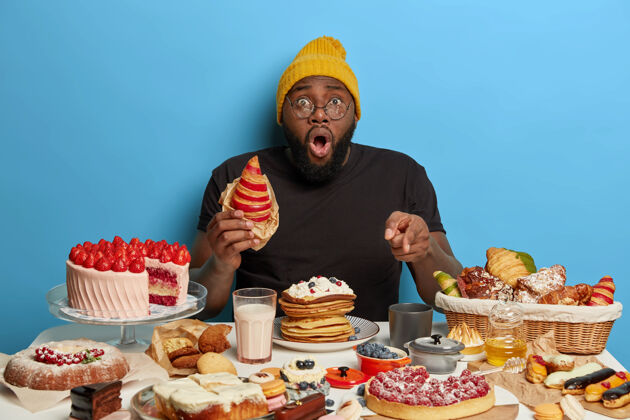 帽子惊呆了的黑人吃着美味的羊角面包 指着桌上满满的甜点 戴着帽子和t恤 在蓝色的背景下摆姿势惊人煎饼蛋糕