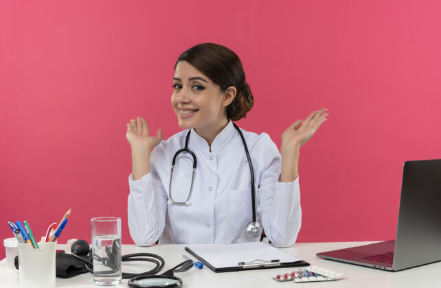 表演微笑着的年轻女医生穿着医用长袍和听诊器坐在办公桌旁 手里拿着医疗工具和笔记本电脑 两手空空的 被隔离在粉红色的墙上长袍笔记本电脑微笑