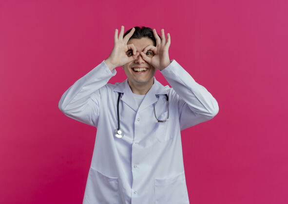 手势微笑着的年轻男医生穿着医用长袍和听诊器做着看手势 用手作为望远镜隔离在粉红色的墙上 留有复印空间使用手听诊器