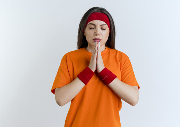 年轻戴着头带和腕带的宁静的年轻运动女性双手合十 闭着眼睛祈祷 隔离在白色墙壁上 留有复制空间在一起女人头带