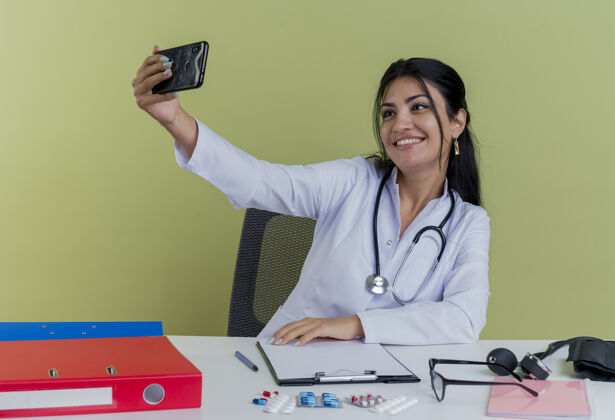 长袍面带微笑的年轻女医生穿着医用长袍 手持听诊器坐在办公桌旁 手持医疗工具 独自在橄榄绿的墙上自拍女性坐着工具
