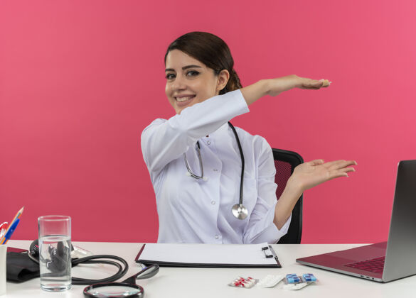 工具微笑的年轻女医生穿着医用长袍和听诊器坐在办公桌旁 带着医疗工具和笔记本电脑 粉红色的墙上显示着孤立的大小微笑表演笔记本电脑