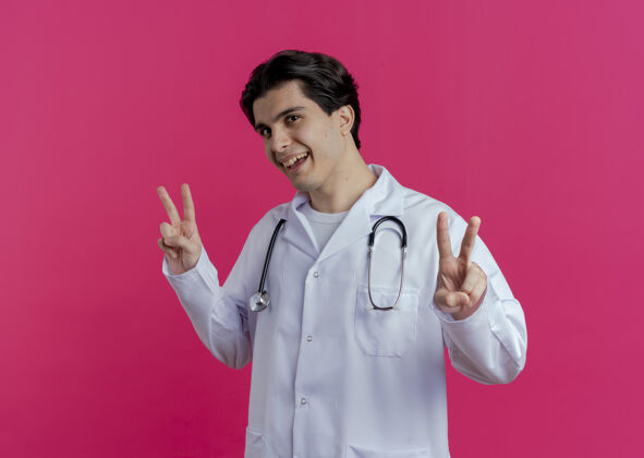 医生令人印象深刻的年轻男医生穿着医用长袍和听诊器做和平标志隔离在粉红色的墙上与复制空间长袍年轻穿