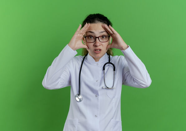 脸年轻的女医生穿着医用长袍 戴着听诊器 戴着眼镜 触摸着隔离在绿墙上有复印空间的脸女看印象