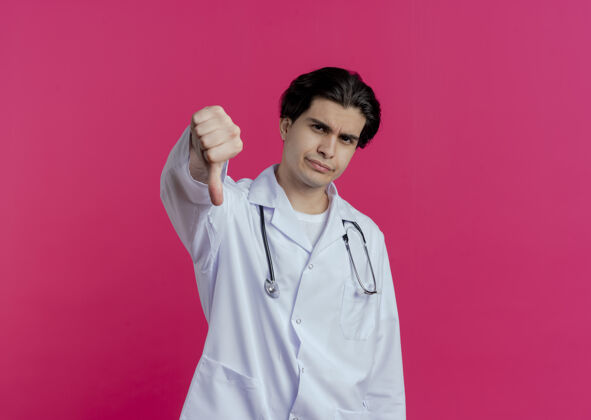 医生皱眉的年轻男医生穿着医用长袍和听诊器 拇指朝下 隔离在粉红色的墙上 留有复印空间皱眉男性听诊器