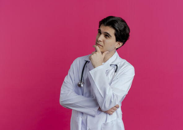 下巴体贴的年轻男性医生 穿着医用长袍 戴着听诊器 看着侧面接触的下巴 隔离在粉红色的墙上 留着复印空间壁板触摸医生
