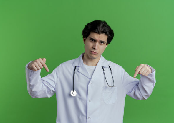 长袍皱眉的年轻男医生穿着医用长袍 听诊器指着隔离在绿色墙上的墙医生指向听诊器