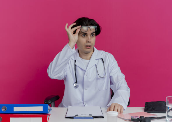 男性年轻的男医生穿着医用长袍 戴着听诊器 戴着眼镜 坐在办公桌旁 拿着医疗工具 看着粉红色墙上的眼镜医生坐着印象