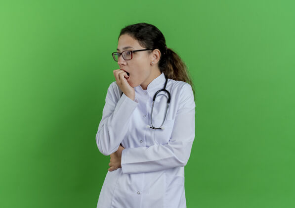 看焦急的年轻女医生 穿着医用长袍 戴着听诊器和眼镜 看着被隔离在绿色墙壁上的手指咬着一边的复印空间眼镜壁板长袍