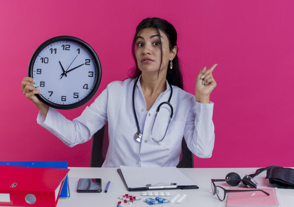 医疗年轻的女医生穿着医用长袍和听诊器坐在办公桌旁 手里拿着医疗工具 手举着时钟 手指被隔离在粉红色的墙上年轻手指坐着