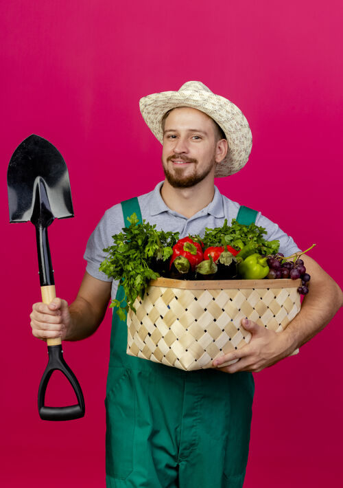 蔬菜高兴的年轻英俊的斯拉夫园丁穿着制服 戴着帽子 手里拿着一篮子蔬菜和铁锹看着请制服帅哥