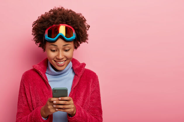高兴活跃的女性滑雪者或滑雪板运动员高兴地看着隔离在粉红色墙上的手机乐观新鲜肖像