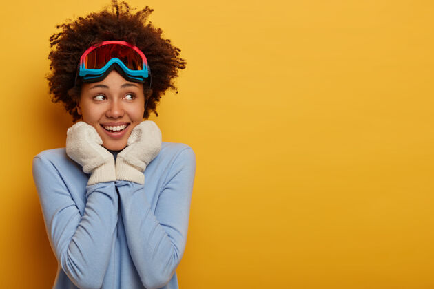 爱好滑雪胜地和滑雪板快乐微笑的黑皮肤女人戴着白手套 戴着滑雪镜和蓝色高领毛衣 站在黄色的背景上运动员活动手套