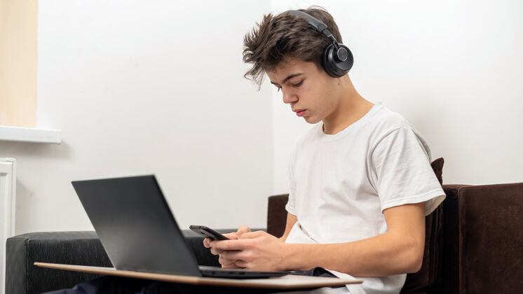 室内十几岁的男孩正在家里用带耳机的智能手机 膝上型电脑严肃而专注的脸控制台青少年笔记本电脑