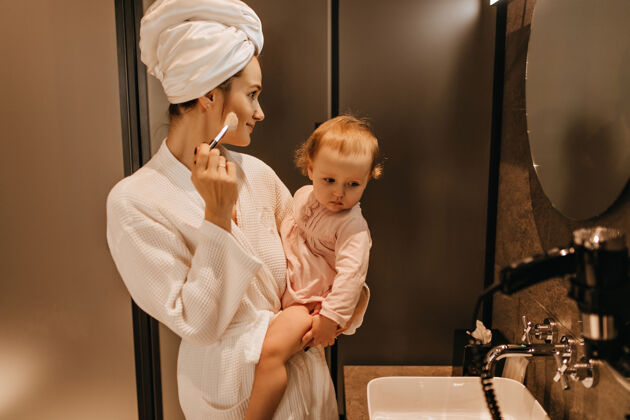 化妆品穿着浴衣的年轻女子抱着金发碧眼的女儿 自己化妆 看着浴室的镜子长袍毛巾刷子