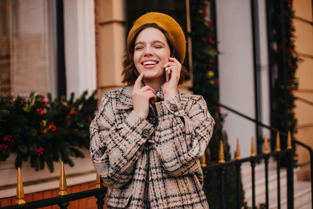 人戴着橙色贝雷帽和外套的积极的女人对着城墙边打电话边笑城市帽子巴黎
