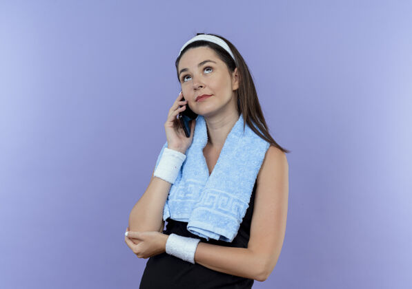 看戴着头巾脖子上挂着毛巾的年轻健身女士站在蓝色的墙上讲着手机抬头思考脖子交谈周围