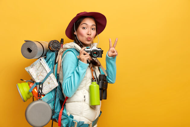 韩国相当活跃的背包客作出胜利的姿态 保持嘴唇圆 持有复古相机 站在旅行袋 在旅行中拍照 隔离在黄色背景青少年休闲旅游