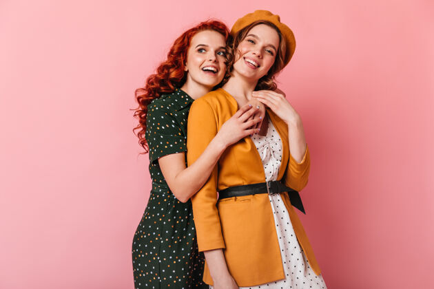 拥抱放松的朋友拥抱在粉红色的背景工作室拍摄的两个可爱的女孩看着相机卷发姜时尚
