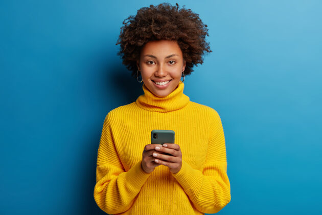 互联网可爱的黑皮肤成年女子穿着黄色套头衫用手机带着幸福的表情打字社交小玩意